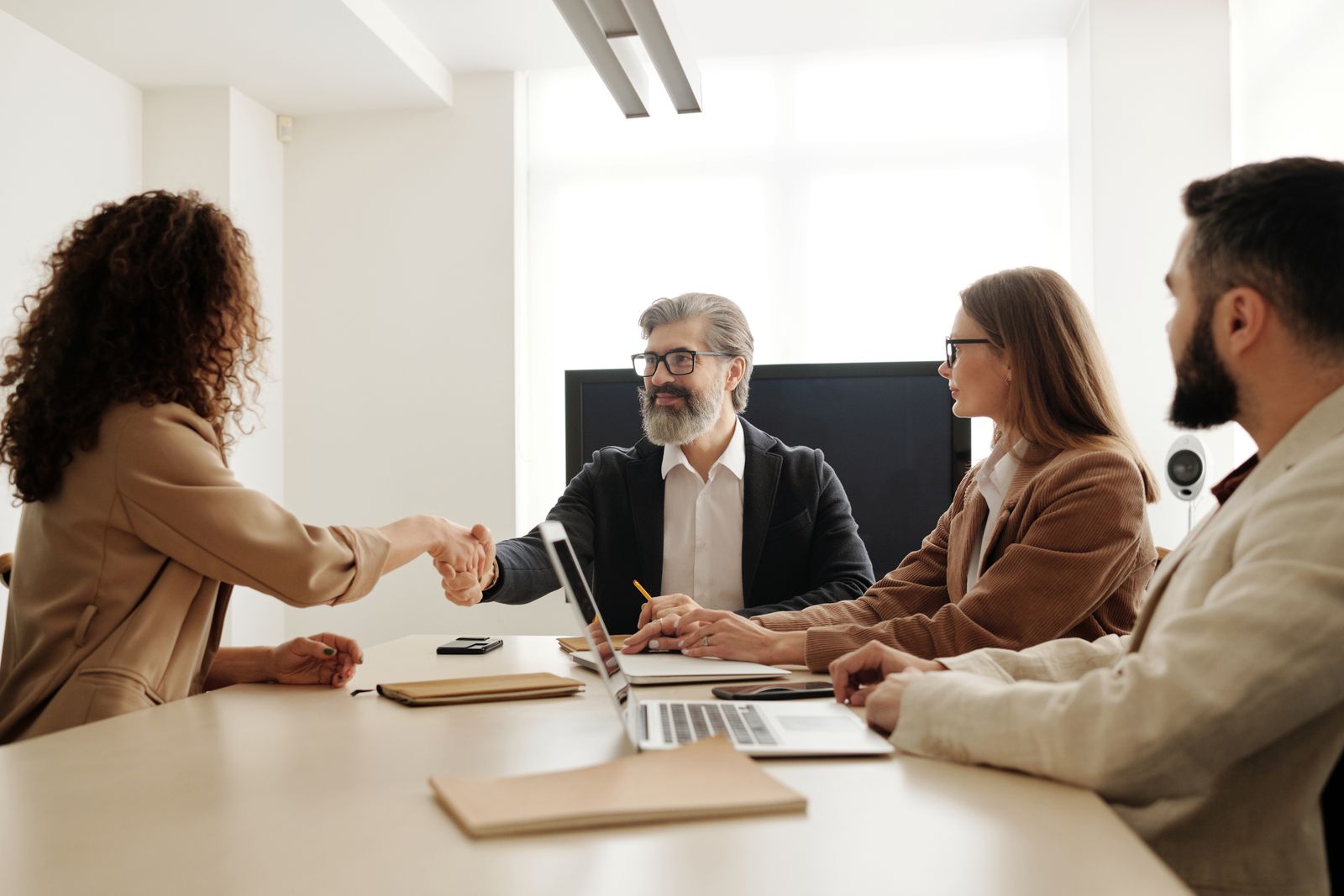 Quatro pessoas em volta de uma mesa de reunião; uma mulher e um homem de barba se cumprimentam enquanto dois colegas observam, com notebooks sobre a mesa.