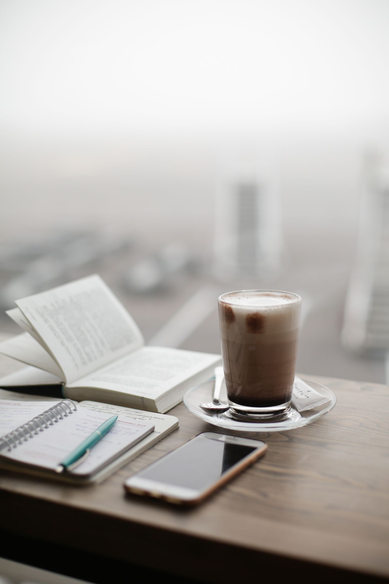 Um copo de café com leite, um livro aberto, um caderno com caneta e um celular sobre uma mesa de madeira junto a uma janela.