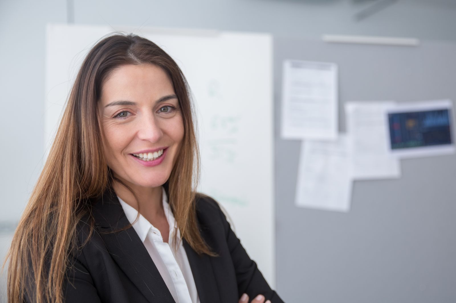 Uma mulher sorridente de blazer preto em um escritório, com um quadro branco e papéis fixados ao fundo.